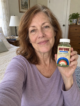 Woman in a lilac top holding UNFUNK, in her bedroom